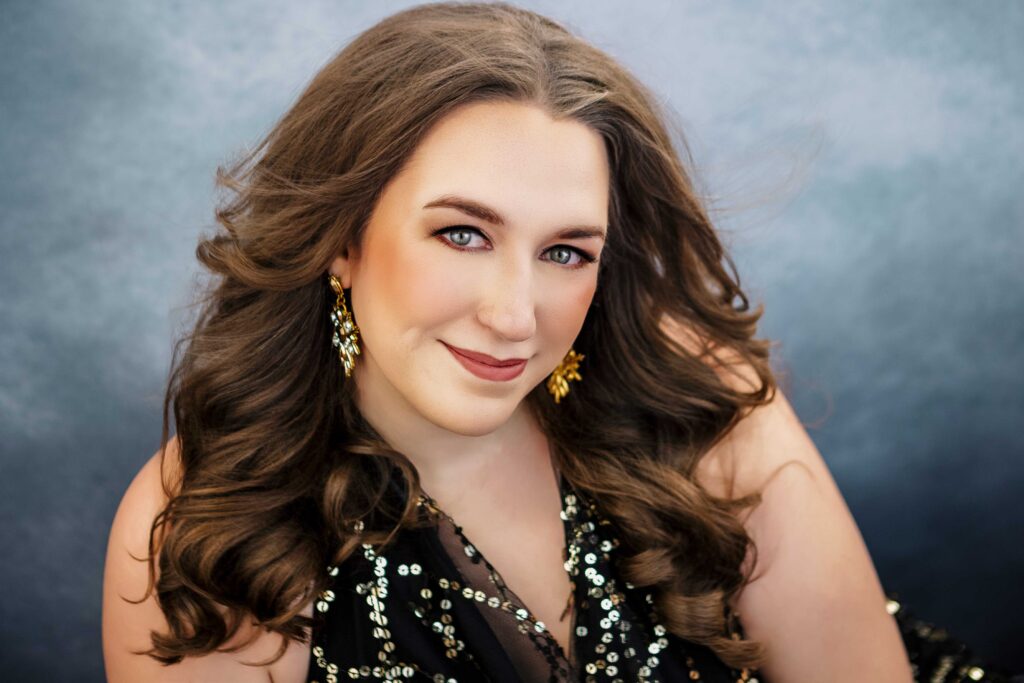 A woman with long, wavy brown hair smiles softly at the camera. She is wearing gold earrings and a black top decorated with sequins, sitting in front of a softly blurred gray background.