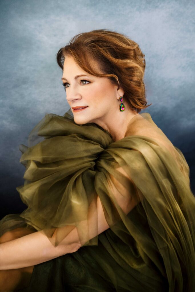 A woman with wavy brown hair wearing a sheer, olive-green gown and colorful earrings poses gracefully against a soft, blue and gray gradient background, looking thoughtfully to the side.