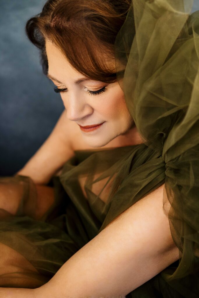 A woman with light skin and reddish-brown hair sits with her eyes closed, wearing a sheer, olive-green tulle dress. She has soft makeup and a peaceful expression. The image is softly lit, creating an elegant, serene mood.