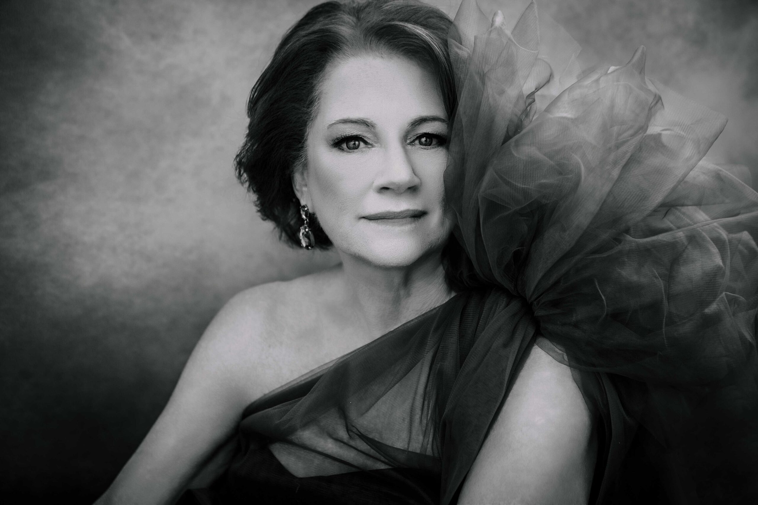Frederick Maryland high-end portrait photography showing a woman stoically looking at camera as she sits in front of gradient background and wears a tulle gown that has a poofy shoulder. The image is black and white.
