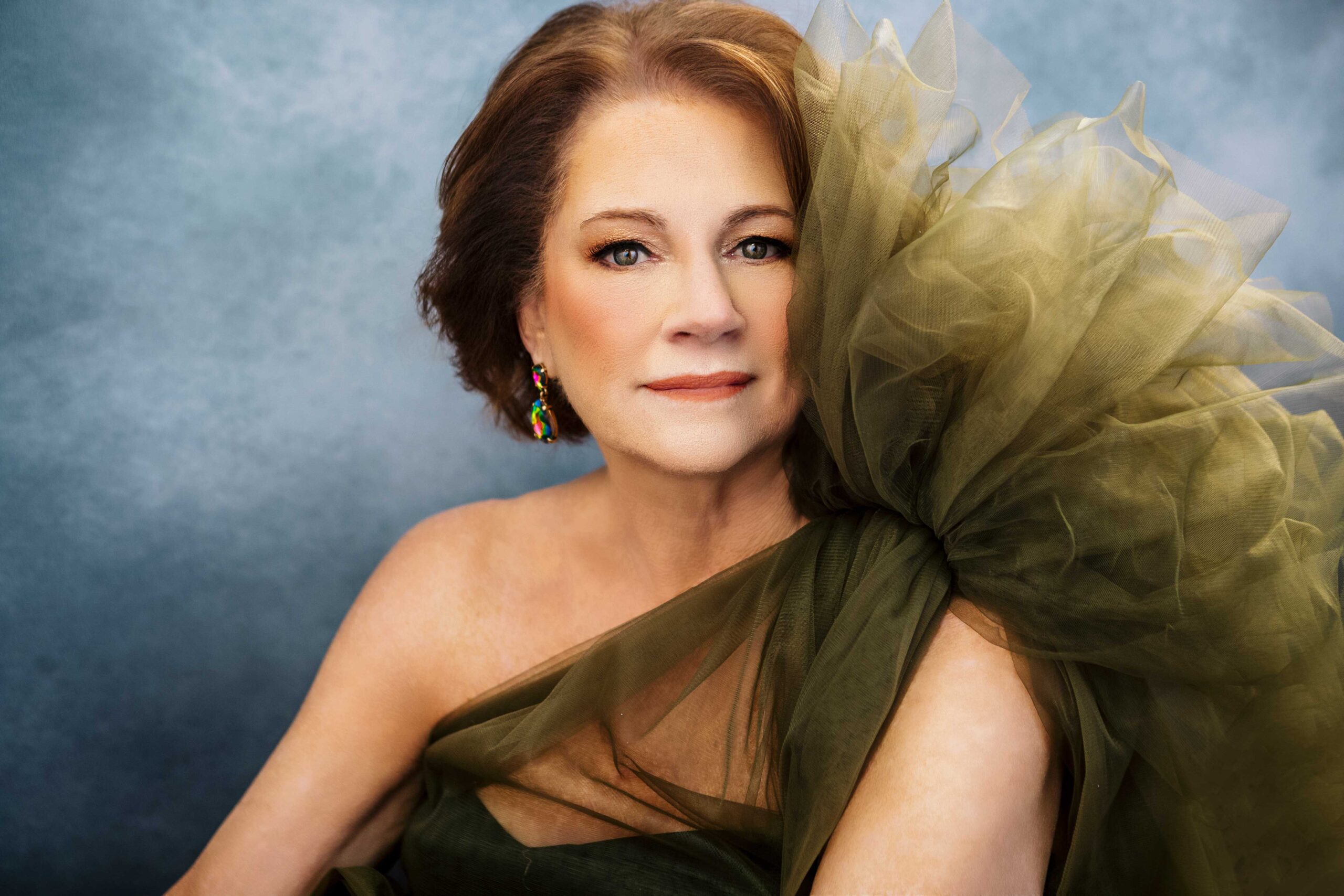 A woman with short dark hair wears a dramatic, tulle-ruffled dress and earrings, gazing confidently at the camera against a softly blurred background. She has colorful drop earrings and sits against a soft blue-gray background, looking slightly to her left