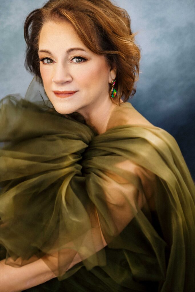 A woman with wavy auburn hair and light makeup wears a sheer, olive green gown with ruffled tulle. She has colorful drop earrings and sits against a soft blue-gray background, looking slightly to her left and smiling gently.