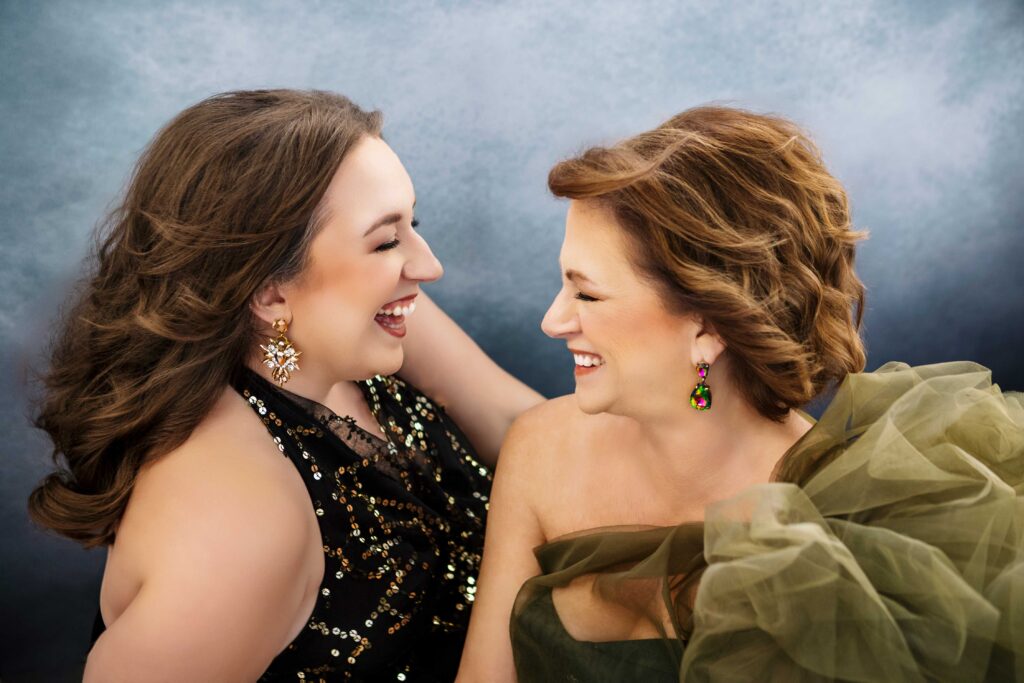 Two women in elegant, olive-green gowns laugh with each other sitting side-by-side surrounded by flowing tulle fabric against a soft blue and gray background.