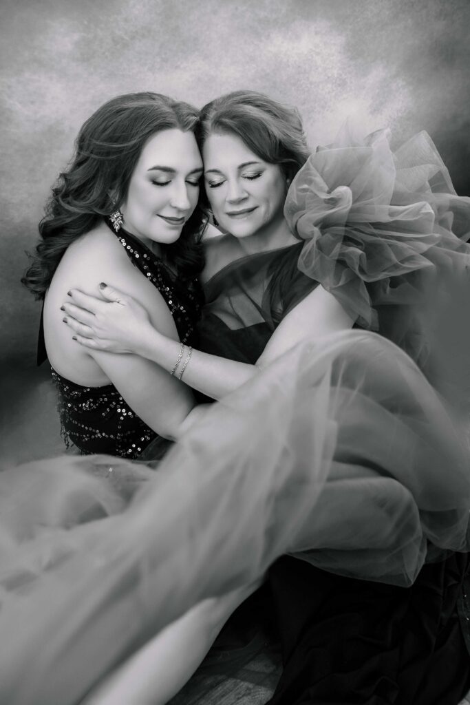 Two women in elegant, gowns smile with their eyes closed and embrace each other, surrounded by flowing tulle fabric against a softly blurred background.