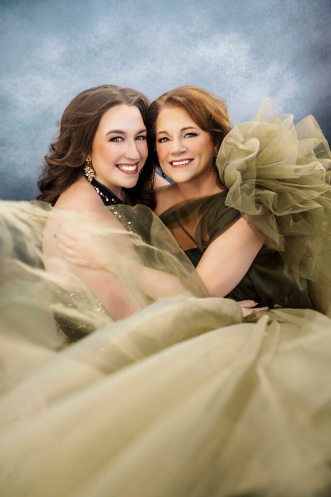 Two women in elegant, olive-green gowns smile and embrace each other, surrounded by flowing tulle fabric against a soft blue and gray background celebrating motherhood together