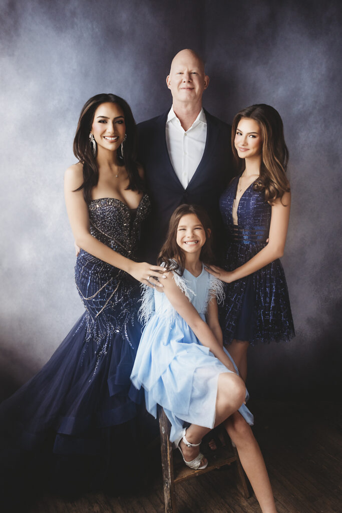 A family of four poses together in formal attire against a dark backdrop. A woman in a navy gown, a man in a suit, and two girls in blue dresses smile warmly, with one girl sitting on a stool in the front are shot by Maryland's best portrait photographer