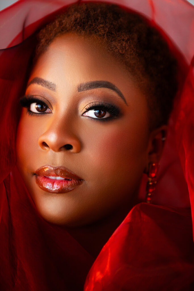 A black woman with short curly hair and luminous makeup gazes forward, surrounded by sheer red fabric. Her expression is calm and confident, with glossy lips and bold eye makeup highlighted by warm lighting in Frederick high-end portrait photography studio