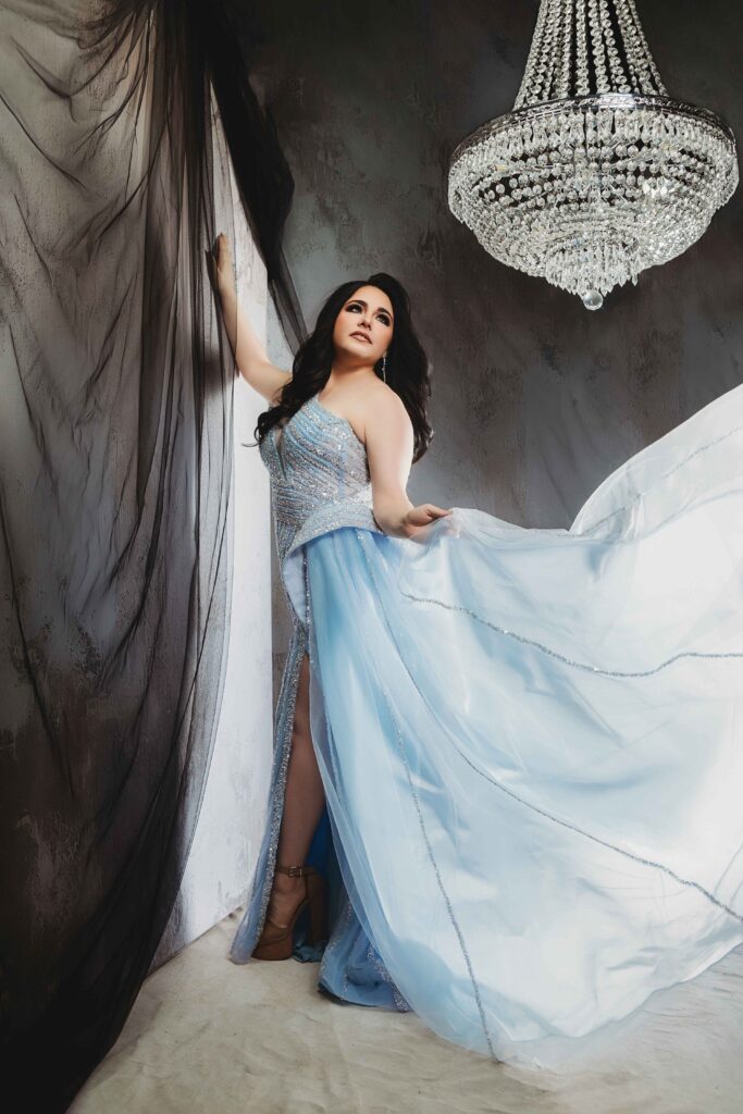 A woman in a flowing light blue gown poses gracefully beside sheer black curtains, holding one side of her dress out. A sparkling crystal chandelier hangs above her against a dark, elegant background.