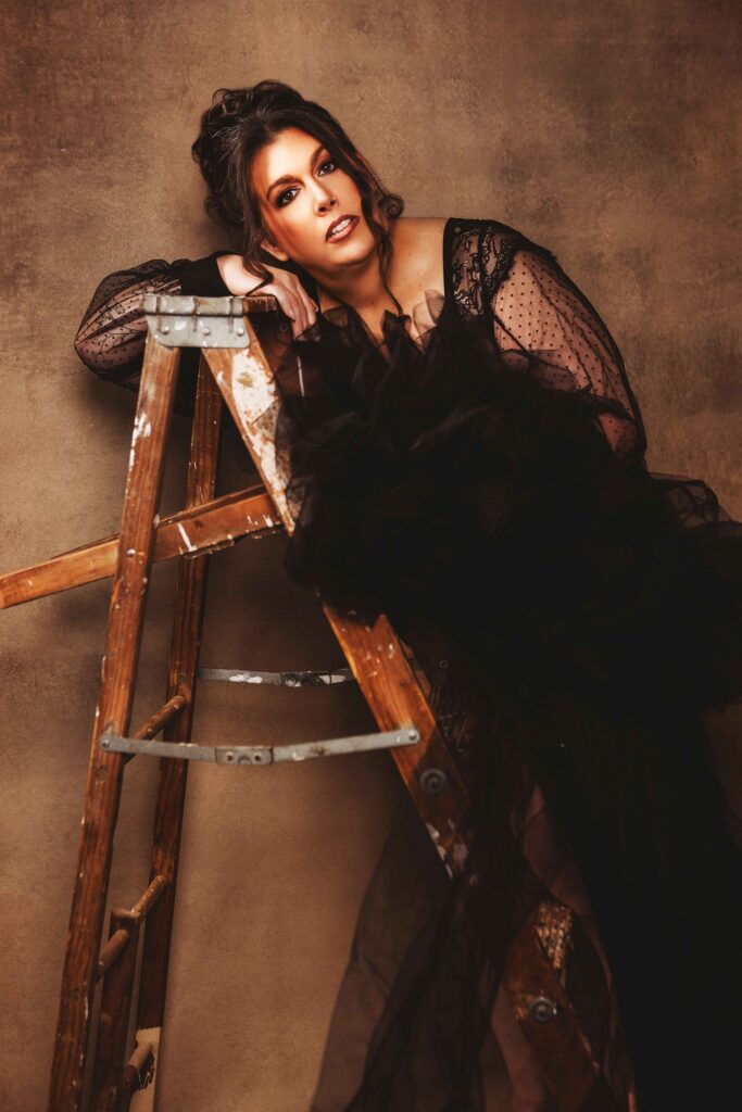 Woman poses with a ladder in front of a brown textured background and wearing a black lace dress for her fine art portrait in baltimore maryland