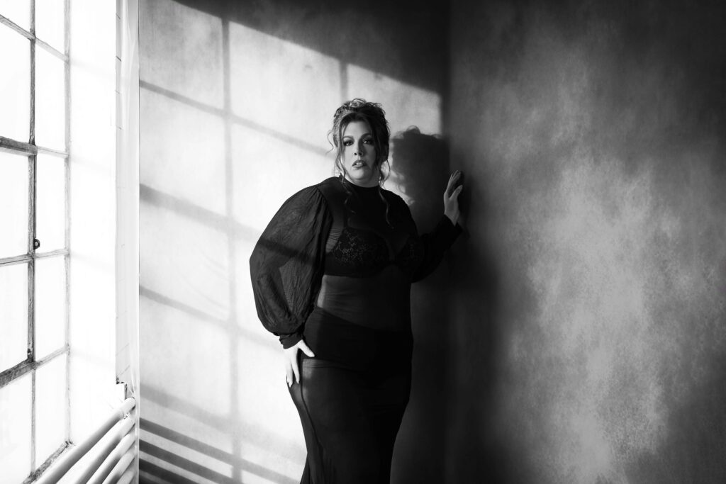 ICON black and white portrait of a woman standing in front of a dark canvas background that has harsh window light on it in frederick maryland