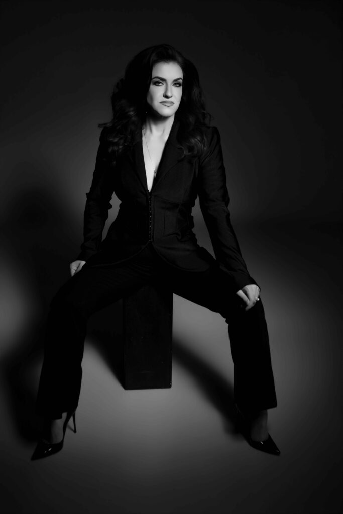 A black and white image of woman wearing a black power suit sits on a back box on a dark background posing strongly for her luxury photo session in Maryland