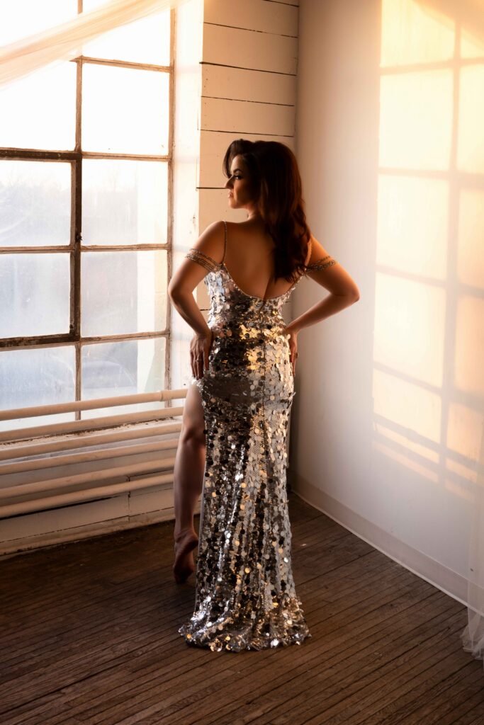 icon portrait of a woman wearing a shiny silver gown in front of a factory window at golden hour with harsh warm light hitting her in aloft studio in maryland